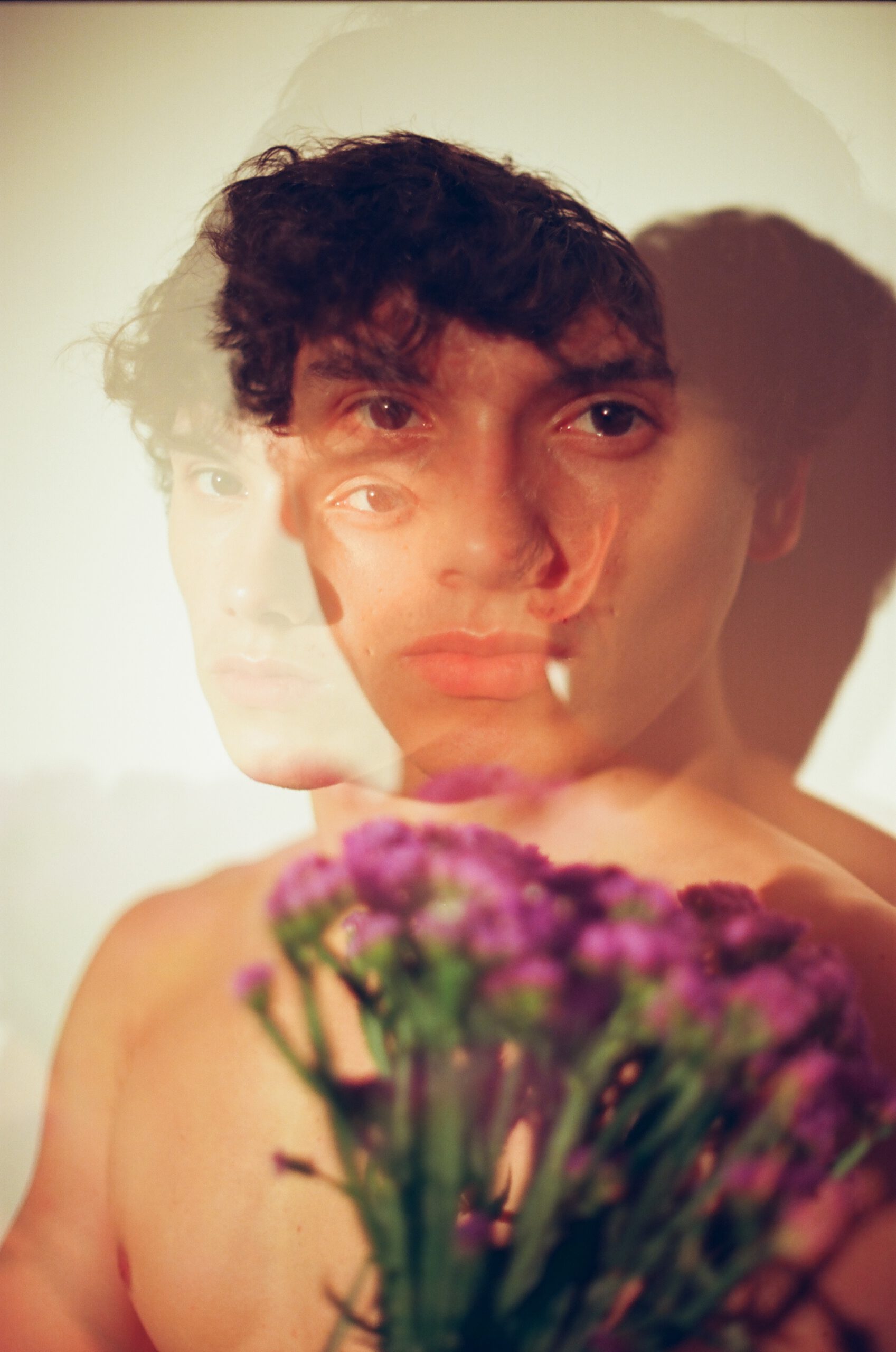 Analog color photograph of a bare male torso holding purple flowers, exploring masculinity, tenderness, desire and vulnerability.